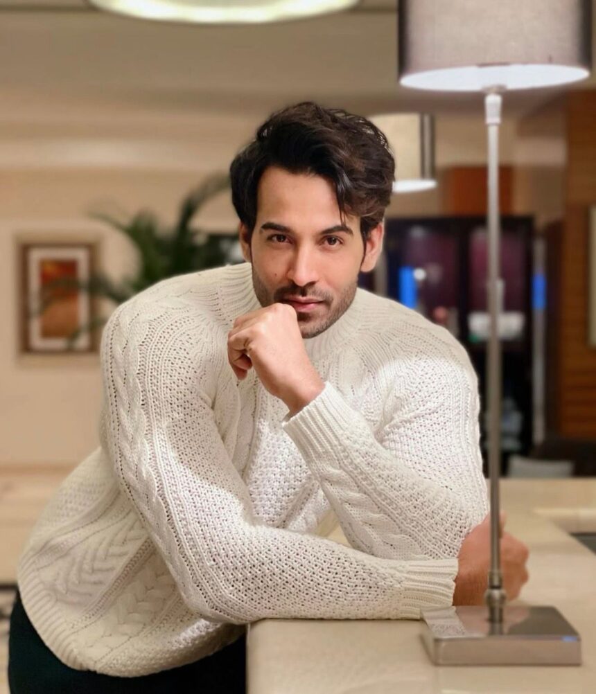 A man posing in a white knitted sweater indoors, seated near a table with a calm and stylish look.