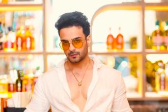 स्टाइलिश अंदाज़ में व्हाइट आउटफिट और ऑरेंज सनग्लासेस पहने अभिनेता, बार काउंटर के सामने पोज़ देते हुए।