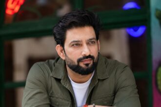 Bearded male celebrity in an olive green jacket posing for a photo
