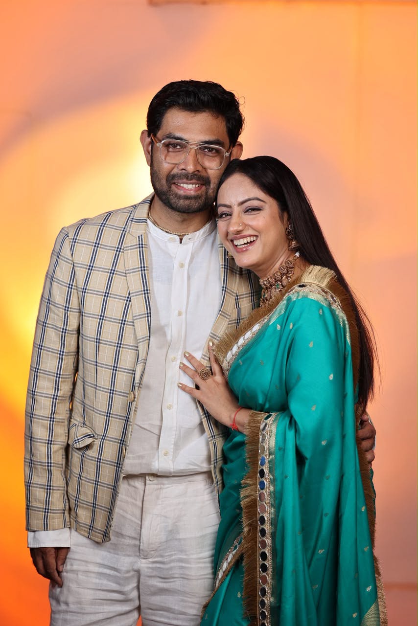 Smiling man and woman posing together, with the woman wearing a turquoise saree and the man in a checked blazer