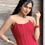 Elegant woman wearing a vibrant red strapless corset-style dress with gold statement jewelry and bracelets, posing against a textured stone wall.