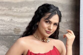 Elegant woman wearing a vibrant red strapless corset-style dress with gold statement jewelry and bracelets, posing against a textured stone wall.