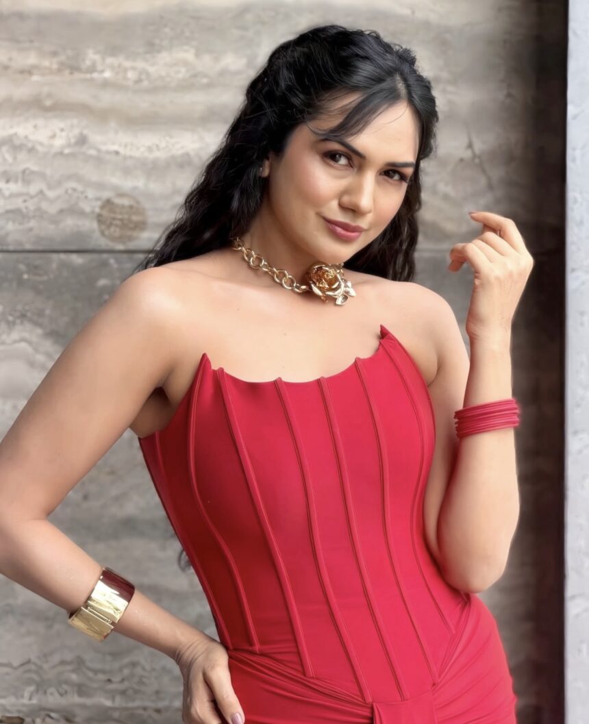 Elegant woman wearing a vibrant red strapless corset-style dress with gold statement jewelry and bracelets, posing against a textured stone wall.